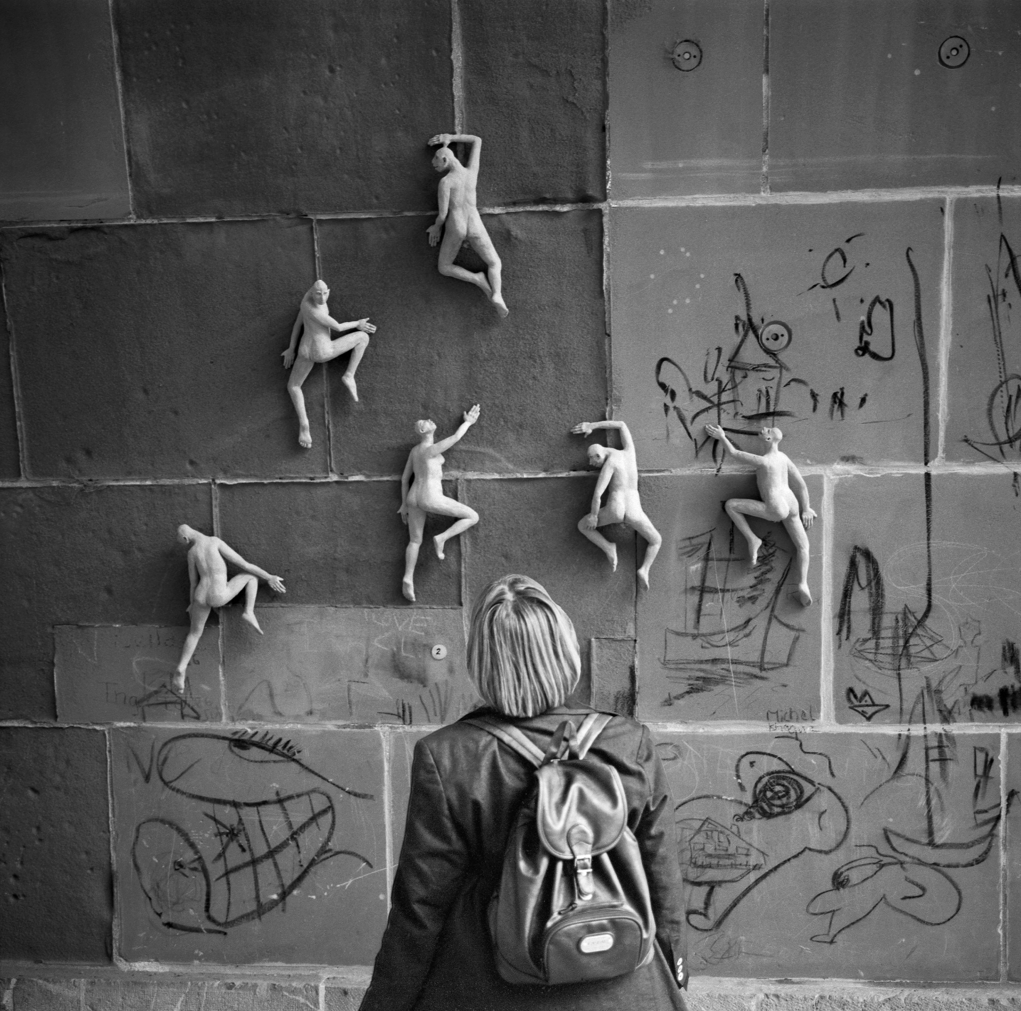 Eine Frau, von hinten gesehen, betrachtet bei einer Vernissage in Bern kleine menschliche Tonfiguren, die an einer mit Zeichnungen bedeckten Wand zu klettern scheinen.